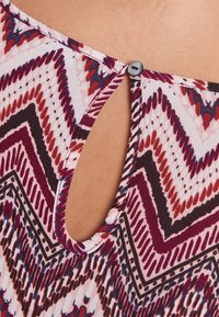 Top de tela con patrón multicolor que presenta un escote con forma de llave y un pequeño cierre de botón en la parte posterior, con diseños en zigzag en rojo, marrón y azul.
