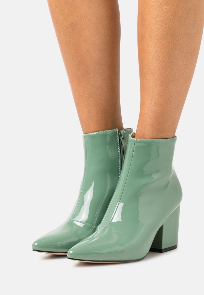 Botas de tobillo de charol verde menta brillante con puntas afiladas, tacones cuadrados y cremalleras laterales, llevadas sobre piernas desnudas contra un fondo blanco.