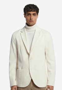 Weißes Cord-Blazer mit Reverskragen, zwei Fronttaschen und einer einzelnen Knopfverschluss, getragen über einem hellen Rollkragensweater.