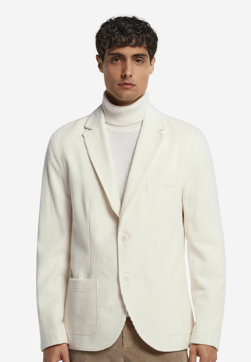 Weißes Cord-Blazer mit Reverskragen, zwei Fronttaschen und einer einzelnen Knopfverschluss, getragen über einem hellen Rollkragensweater.
