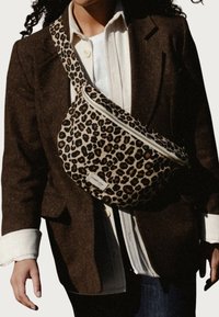 Sac à dos en cuir imprimé léopard, fabriqué en tissu doux, avec une fermeture éclair et une petite étiquette de logo. Porté en bandoulière sur un blazer marron.