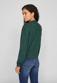 Groene trui met een gestructureerde afwerking, geribbelde manchetten en zoom. Heeft pofmouwen en een hoge halslijn, gecombineerd met blauwe jeans.