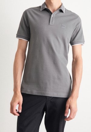Mann trägt ein graues kurzärmeliges Poloshirt mit weißer Einfassung an Ärmeln und Kragen, kombiniert mit schwarzen Hosen.