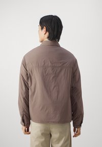 Brown lightweight jacket with a smooth texture, featuring a collared neckline and buttoned cuffs, worn over beige trousers.