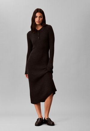 Femme aux longs cheveux foncés portant une robe midi noire côtelée à manches longues avec col et boutons, et des mocassins foncés, debout devant un fond gris.