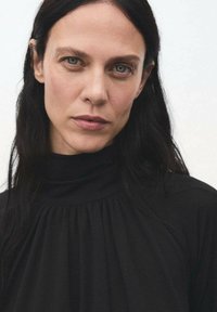 Prenda negra de cuello alto con pliegues verticales, textura suave y diseño minimalista. El sujeto tiene el cabello largo y oscuro y un maquillaje neutro.