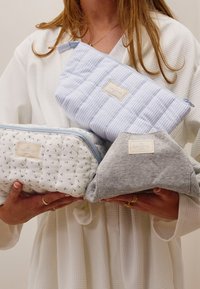 Tre kosmetikväskor i varierande mönster: vit med grönt blommönster, ljusblå med ränder och grått tyg. Alla har dragkedjor och etiketter.