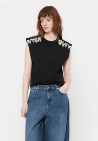 Svart ermeløs topp med skulderdekorasjoner i perle- og krystalldetaljer, kombinert med lyseblå høytlivsjeans. Standard passform.