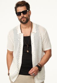 Weiße, gestrickte, kurzärmelige Strickjacke mit offenem Netzmuster, über einem schwarzen Tanktop getragen. Die Accessoires umfassen gestapelte Halsketten und eine Uhr.
