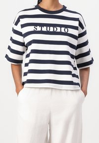 Camiseta de manga corta a rayas en blanco y azul marino, con cuello redondo y texto bordado "STUDIO" en el centro. Diseño de corte relajado.
