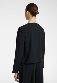 Musta, pitkähihainen takki, jossa on rento leikkaus ja pyöreä kaula-aukko. Materiaali näyttää sileältä ja hieman tekstuurilta. Yksinkertainen, minimalistinen muotoilu.