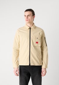 Khaki veekindel jakk, millel on lukuga esiosa, rindade tasku, kus on punane logo, ja mustad tõmblukud. Pikkade varrukatega ja nööbkinnistega.