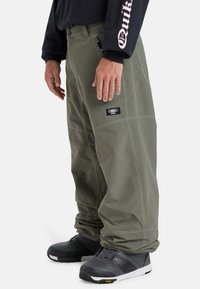 Green cargo pants with stitched accents, side pockets, and a black logo patch, paired with black snow boots. Cotton blend material.