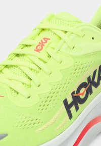 Τρέχον παπούτσι Hoka σε νέον κίτρινο χρώμα με επάνω μέρος από δίχτυ, επενδυμένη γλώσσα, κορδόνια και μαύρο-κόκκινο λογότυπο Hoka στη πλευρά και στη γλώσσα.