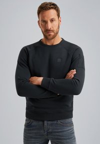 Donkerblauwe sweatshirt met een ronde halslijn, geribbelde manchetten en een gestructureerde logopatch op de borst. Draag met vervaagde blauwe jeans.