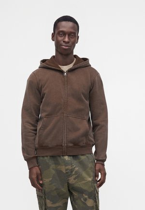 Homme debout devant un fond uni, portant un sweat à capuche zippé marron et un pantalon cargo camouflage vert, regardant devant lui avec une expression neutre.