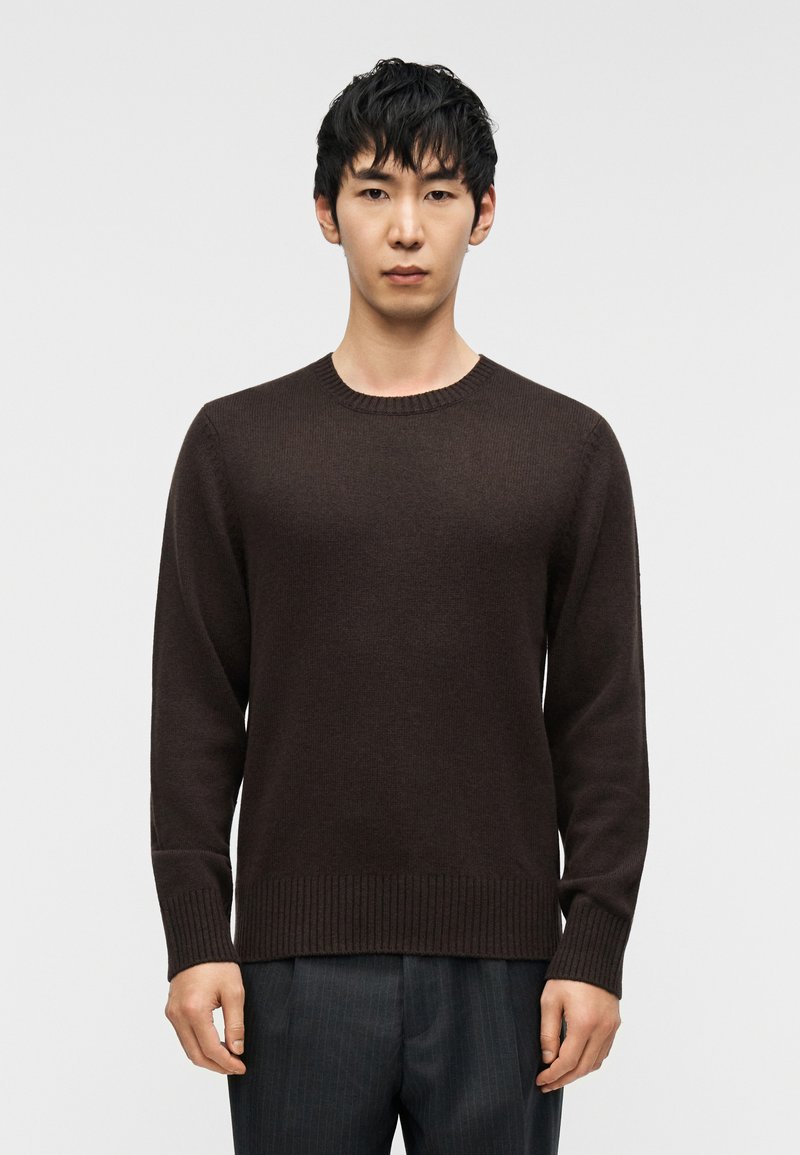 Καφέ πουλόβερ με μακριά μανίκια, ριμπ λαιμόκοψη και τελείωμα, κατασκευασμένο από μαλακό υλικό. Φορεμένο πάνω από σκούρο παντελόνι με ρίγες.