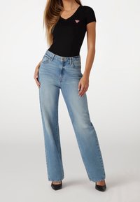 Zwarte korte mouwen v-hals top met een klein rood driehoeklogo, gecombineerd met lichtblauwe hoge taille wijdvallende jeans en zwarte hakken.
