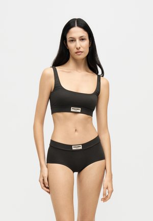 Femme aux longs cheveux foncés portant un crop top noir et des sous-vêtements assortis avec des étiquettes de marque sur un fond clair uni.