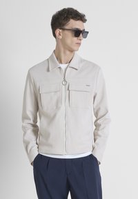 Chaqueta de color beige claro con cremallera, dos bolsillos en el pecho, un tirador en forma de anillo al frente y cuello con solapa, llevada sobre una camiseta blanca y pantalones azul marino.