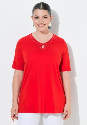 Smilende kvinde med mørkt hår trukket tilbage, iført store hængende øreringe, en rød top med korte ærmer og rhinsten samt hvide bukser.