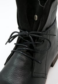 Bottine en cuir noir avec bout arrondi, lacets noirs noués, détails cousus et un petit bouton métallique près du haut de la languette.