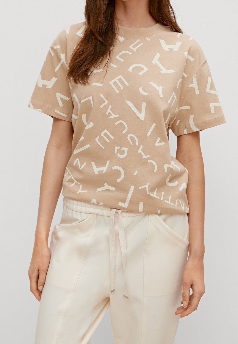 Beige bomulds t-shirt med oversized pasform og store kontrasterende hvide bogstaver over stoffet. Parret med lette, løstsiddende bukser.