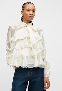 Νεαρή γυναίκα με πλεξούδες φοράει διάφανη ελεφαντόδοντη μπλούζα με στρωτά βολάν και μεγάλο φιόγκο, συνδυασμένη με σκούρο μπλε τζιν.