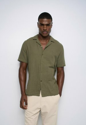 Homme aux cheveux courts portant une chemise olive à manches courtes boutonnée et un pantalon beige sur un fond clair uni.