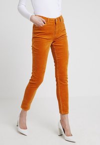 Pomarańczowe spodnie z korduroy o slim fit i wysokim stanie, z wyraźnymi liniami tekstury. Połączone z białymi butami na wysokim obcasie.