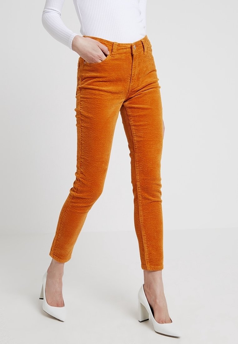 Pomarańczowe spodnie z korduroy o slim fit i wysokim stanie, z wyraźnymi liniami tekstury. Połączone z białymi butami na wysokim obcasie.