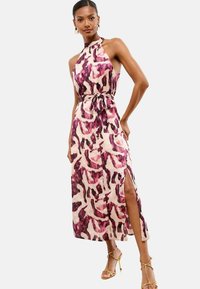 Donna che indossa un abito midi senza maniche, con collo all'americana, in un motivo astratto viola e beige, cintura annodata, spacco laterale e sandali con tacco alto dorati.