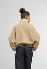 Pullover in pile beige con colletto alto, vestibilità comoda e tessuto testurizzato. Abbinato a jeans in denim scuro con tasche posteriori.