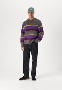 Szürke gyapjú pulóver lila, bordó és sárga csíkokkal, fekete farmernadrággal és fekete sportcipővel, ami gumi talppal rendelkezik. Zöld sapka.