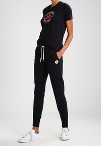 Svart t-shirt med en röd och vit Converse-logotyp, i kombination med svarta joggers och vita sneakers. Enkel design med avslappnad passform.