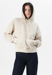 Hoodie cropped beige avec une poche avant, capuche à cordon de serrage et poignets côtelés. Porté sur un jean bleu foncé, mettant en valeur une coupe décontractée et un design confortable.