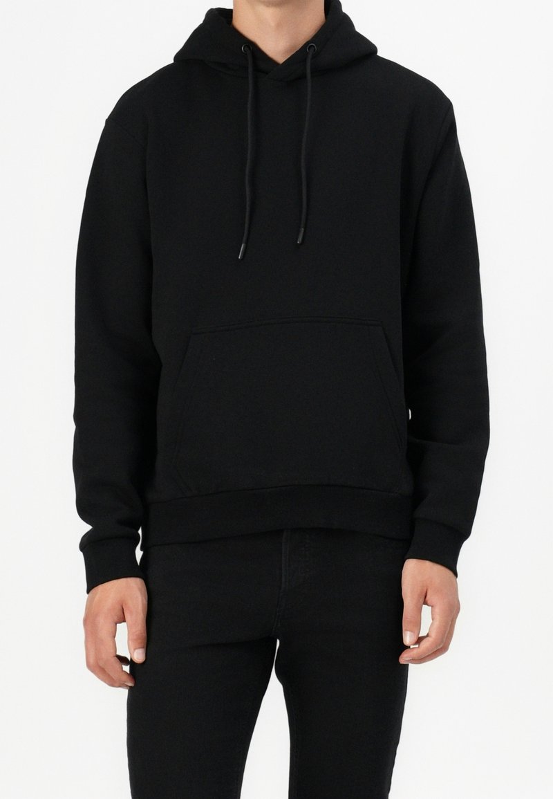 Homme portant un sweat à capuche noir uni avec poche avant et cordons, associé à un pantalon noir, sur un fond blanc.