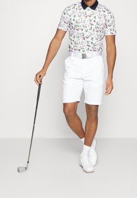 Polo à manches courtes avec motif floral en rose et vert, associé à un short blanc et une ceinture argentée. Club de golf tenu dans la main droite.