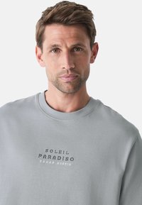 Šviesiai pilkas sportinis megztinis su apvaliu kaklu ir raštuotais rankogaliais, ant priekinės dalies užrašas "SOLEIL PARADISO TEXAS AUSTIN".