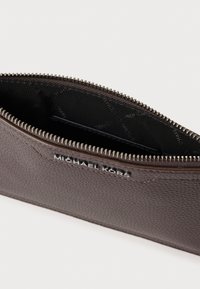 Barna bőrből készült cipzáras tok, amely texturált felülettel, bélelt belsővel és ezüst színű fém kiegészítőkkel rendelkezik, a külső felületén dombornyomott logóval.