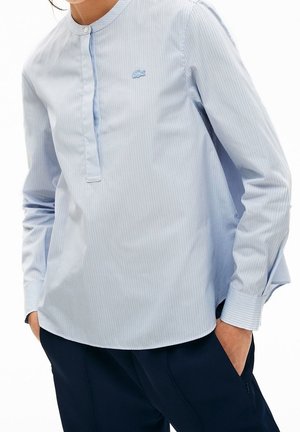 Vrouw draagt een lichtblauw overhemd met verticale strepen, lange mouwen en een colbertkraag, met een donkere blauwe broek, handen in de zakken.