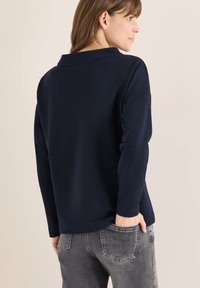 Marineblaues Polohemd mit langen Ärmeln und Kragen, aus weichem Stoff; von hinten gezeigt mit grauer Jeans, wobei die Taschendetails hervorgehoben werden.