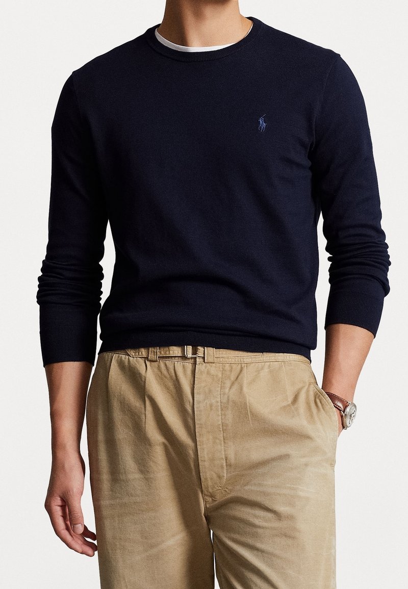 Homme portant un pull à manches longues bleu marine avec un petit logo, un pantalon beige et une montre-bracelet, se tenant debout avec une main dans la poche.