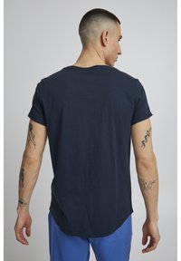 Svart t-skjorte med korte ermer og rund kant, laget av mykt stoff, med rullede ermer og synlige tatoveringer på underarmene.