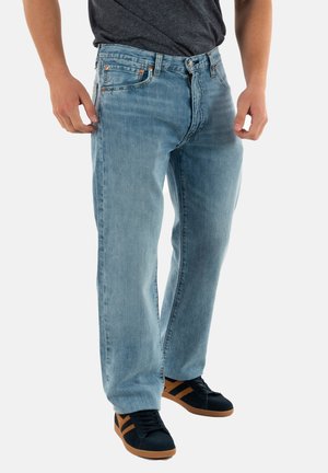 Jeans in denim azzurro chiaro con taglio dritto, design a cinque tasche e componenti in ottone. Indossati con scarpe da ginnastica nere con dettagli marroni.