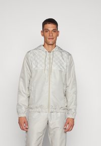 Chaqueta con cremallera de color gris claro y capucha, con un patrón de diamantes texturizado en el pecho y puños elásticos. Tela suave y ligera.