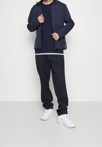 Marinblå jacka med vadderade axlar, fickor med dragkedja, tillsammans med en marinblå tröja med rund halsringning och matchande smalnande byxor. Vita sneakers fulländar looken.