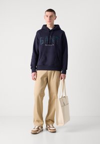 Felpa in cotone blu navy con logo "GANT", pantaloni in cotone beige, e borsa a tracolla beige; sneakers brown e bianche con strisce diagonali.