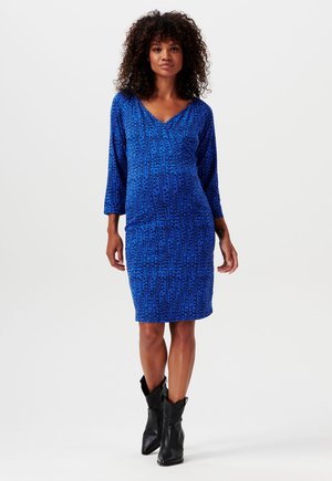 Sininen kuviollinen mekko, jossa on kolmivartiset hihat, V-kaula-aukko, tiukka siluetti ja polvimittainen pituus. Yhdistetty mustiin nilkkasaappaisiin.