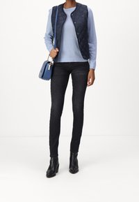 Marineblau gesteppte Weste über einem hellblauen Langarmshirt, kombiniert mit schwarzen engen Jeans und schwarzen Absatz Stiefeletten. Blauer Handtasche mit Hardware-Akzenten.